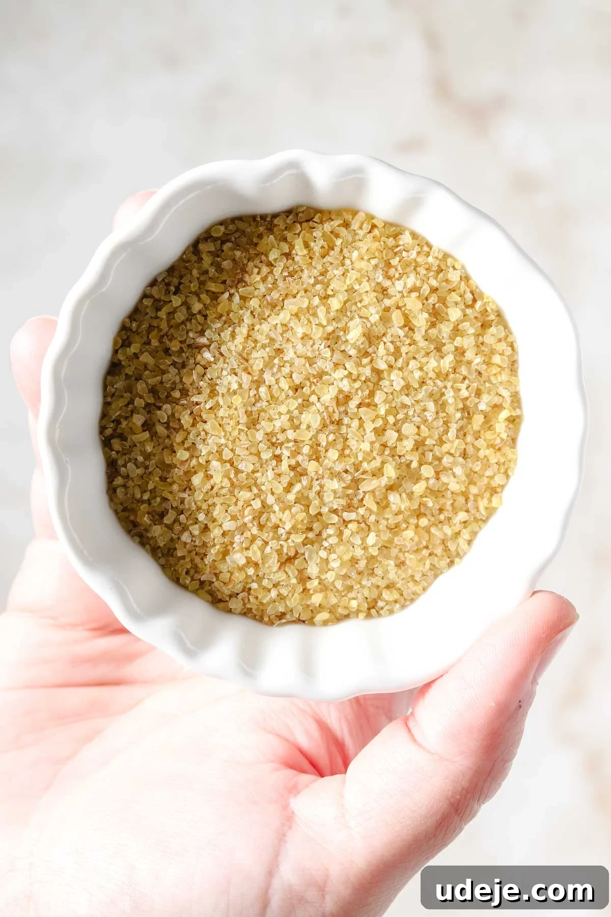 Fast 20-Minute Turkish Bulgur Kisir 3 Unsoaked fine bulgur grains in a white bowl, held gently by a hand against a light marble background, showcasing its characteristic texture before preparation.