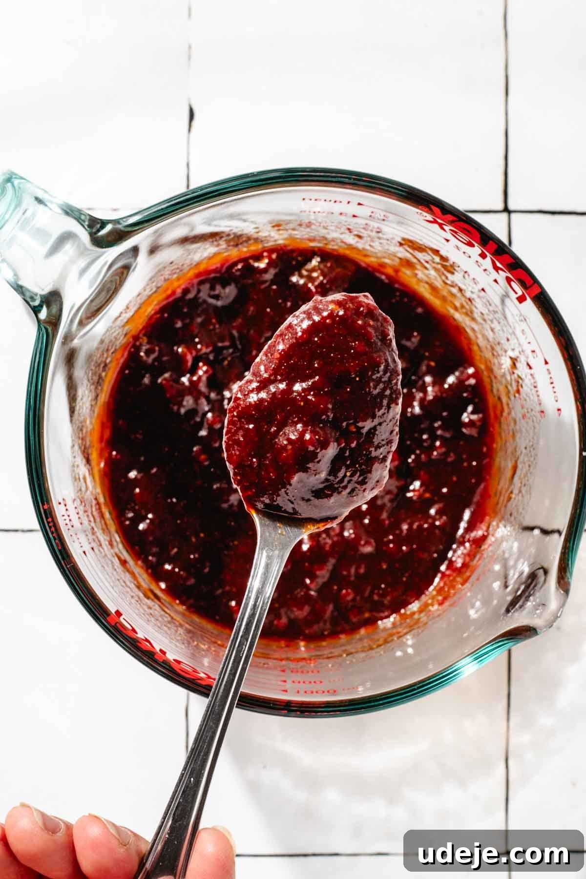 Fast 20-Minute Turkish Bulgur Kisir 9 A spoonful of rich, red Kisir dressing held up from a glass measuring cup, showcasing its thick, paste-like consistency, against a white tiled background.