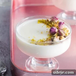 A glass dessert cup filled with creamy Mahalabia, garnished with chopped pistachios and dried rosebuds, sits on a pink tray. Another similar Muhallebi dessert is visible in the background.