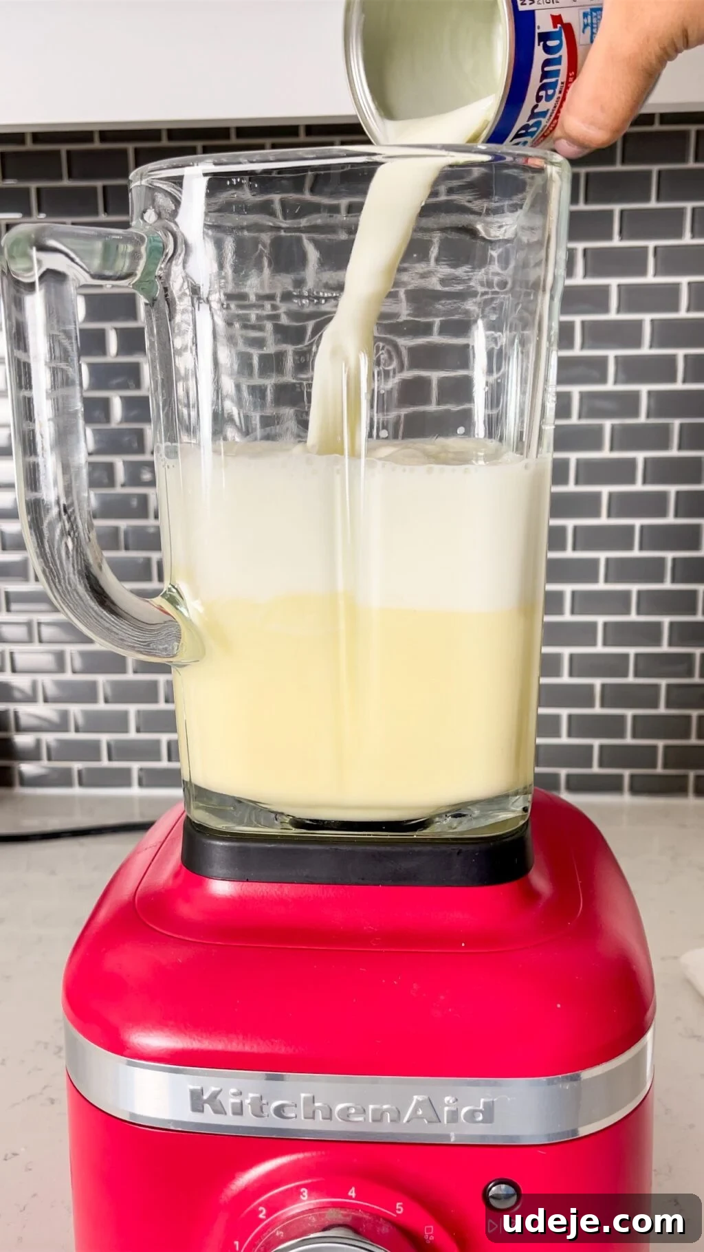 Creamy Condensed Milk Creme Caramel 11 milk pouring into a glass blender jug on a red bottom with a dark grey tiled backsplash