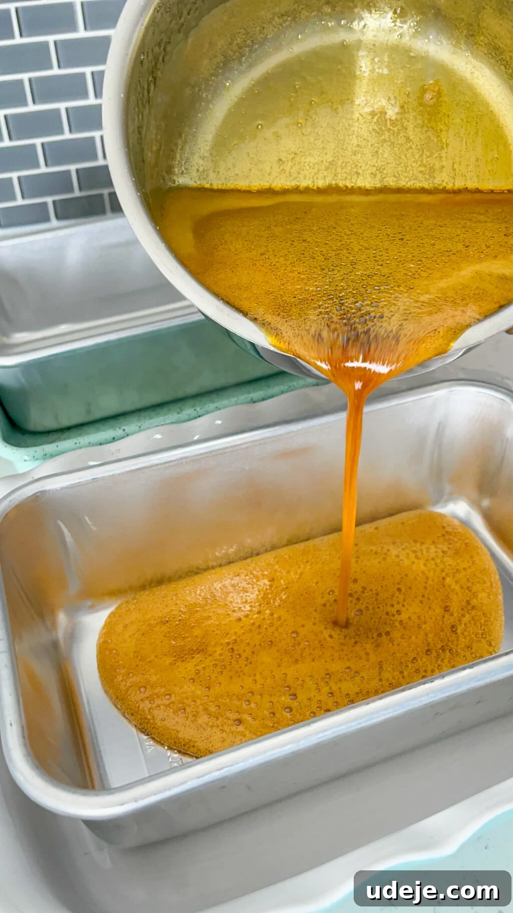 Creamy Condensed Milk Creme Caramel 14 caramel being poured from saucepan into steel loaf tin with a dark grey tiled backsplash
