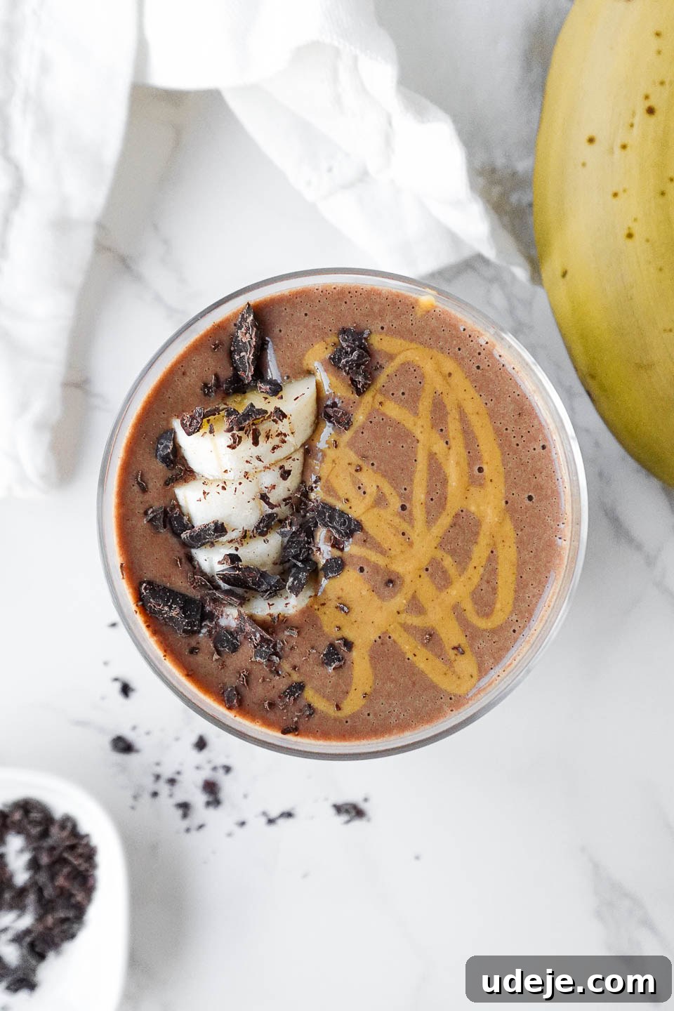 Speedy Chocolate Banana Indulgence 2 A vibrant vegan chocolate banana smoothie, perfectly blended and served in a clear glass. It's elegantly topped with a sprinkle of cacao nibs, fresh banana slices, and a luscious drizzle of peanut butter, highlighting its rich texture and appealing presentation.
