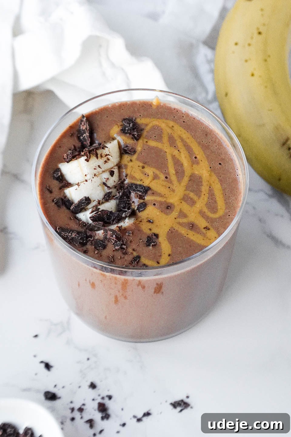 Speedy Chocolate Banana Indulgence 3 A close-up of a vegan chocolate banana smoothie being poured into a tall glass, showcasing its thick, creamy texture. The surface is adorned with a sprinkle of cacao nibs, fresh banana slices, and a delicate swirl of peanut butter, inviting a delicious sip.