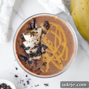A beautifully presented vegan chocolate banana smoothie, garnished with cacao nibs, fresh banana slices, and a rich swirl of peanut butter, captured from an appealing angle.