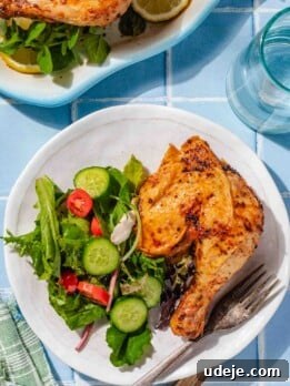 air fried chicken leg on a white plate with a salad and fork with a glass of water and the whole air fried chicken on a white platter on a blue tiled background