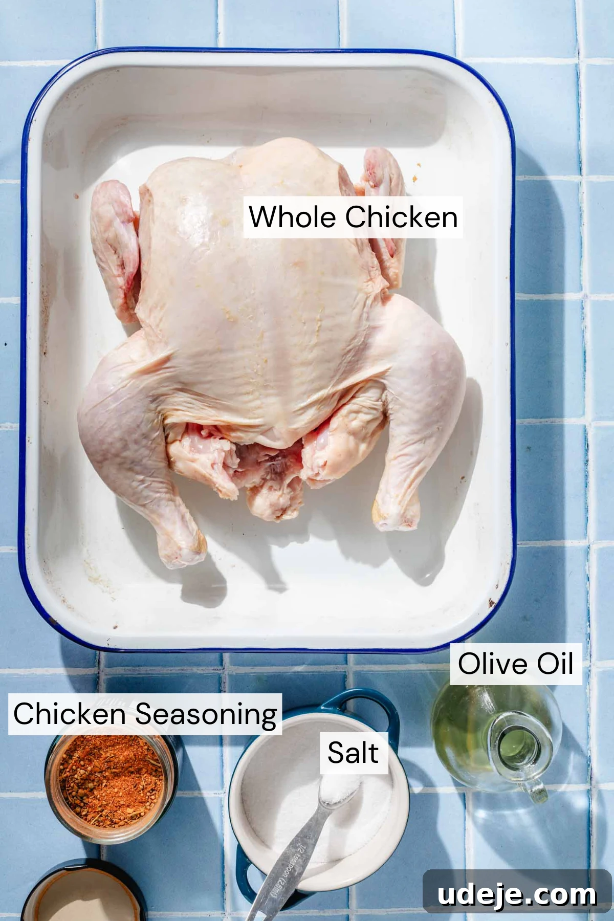 Crispy and Juicy Air Fryer Whole Chicken in 60 Minutes 3 Ingredient shot for Air Fryer Whole Chicken with a whole chicken in a blue rimmed white baking pan, a small pitcher of olive oil, a small jar of chicken seasoning and a small blue pot of salt with a metal measuring spoon on a blue tiled background