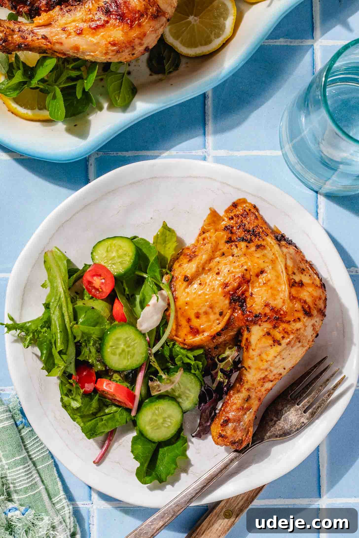 Crispy and Juicy Air Fryer Whole Chicken in 60 Minutes 9 air fried chicken leg on a white plate with a salad and fork with a glass of water and the whole air fried chicken on a white platter on a blue tiled background