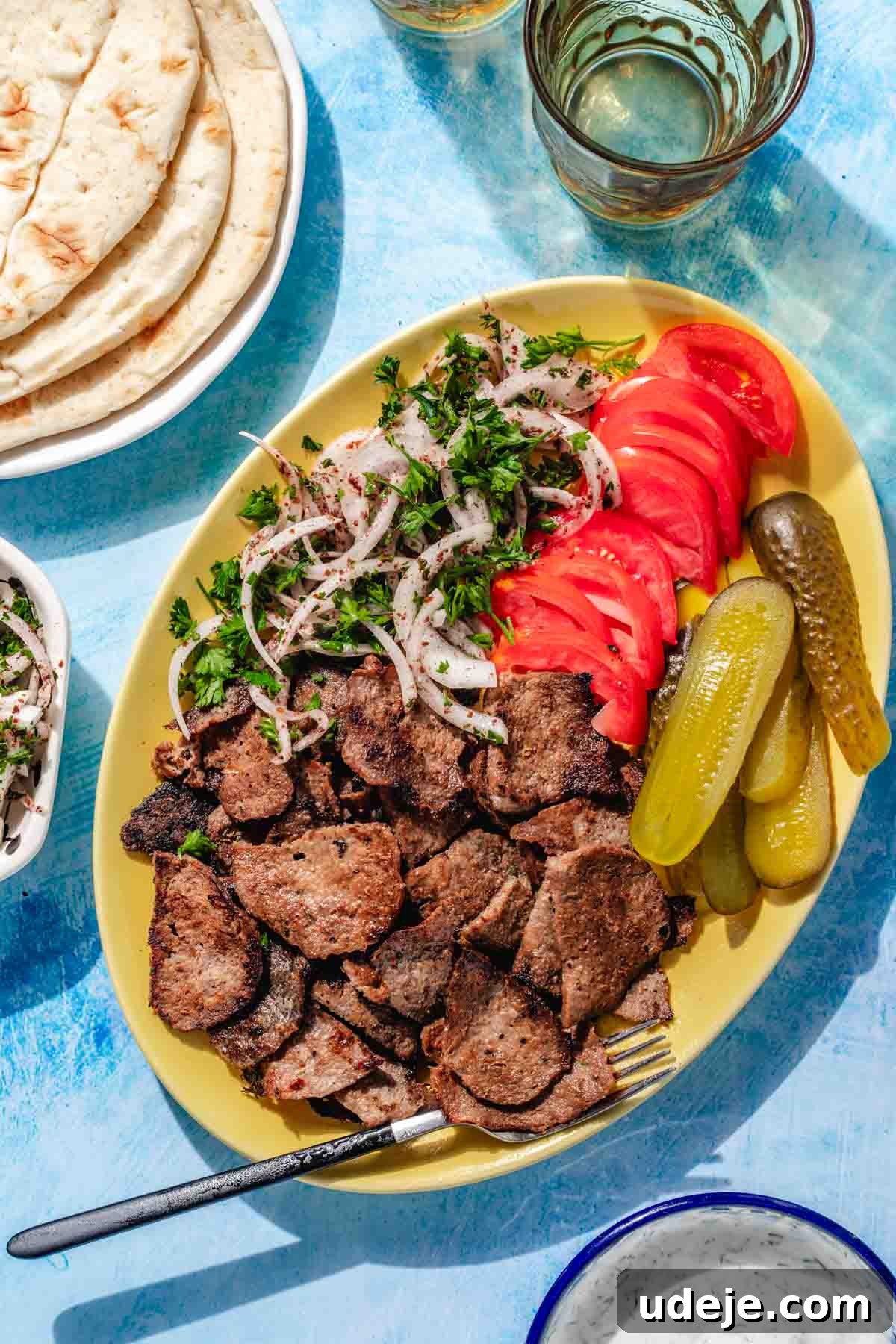 One-Pan Lamb and Beef Doner Kebab 2 A vibrant yellow platter showcasing beautifully cooked doner meat slices, accompanied by fresh sliced red onions, ripe tomato slices, crisp pickled cucumbers, and a fork. In the background, a blue surface with a plate of warm pita bread and a small bowl of classic white doner sauce, alongside a refreshing glass of water.