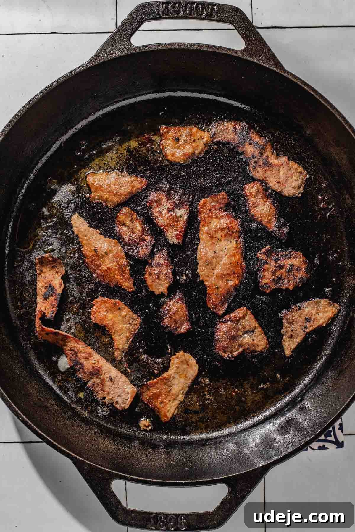 One-Pan Lamb and Beef Doner Kebab 16 Thin slices of doner meat are actively frying in a hot cast iron skillet, creating a sizzling and aromatic scene against a clean white tiled background, developing a golden crust.