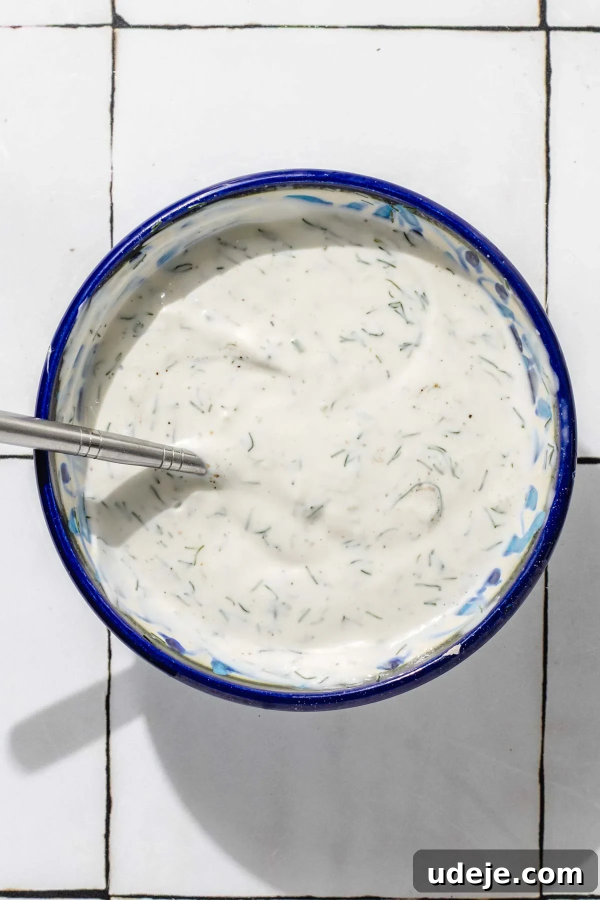 One-Pan Lamb and Beef Doner Kebab 17 A beautifully textured white doner sauce fills a blue patterned bowl, with a metal spoon resting within, positioned elegantly on a white tiled background, ready to be served.