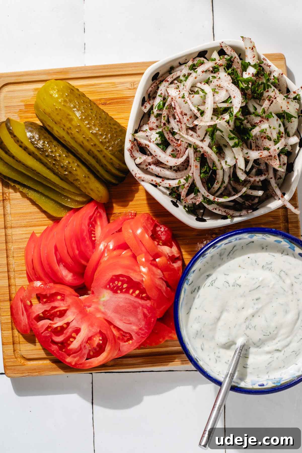 One-Pan Lamb and Beef Doner Kebab 18 A rustic cutting board laden with fresh Doner kebab toppings: bright green sliced pickles, vibrant red sliced tomatoes, and a white bowl filled with thinly sliced onions and fragrant herbs. A blue patterned bowl holds the creamy white doner sauce with a metal spoon, all arranged enticingly on a white tiled background.