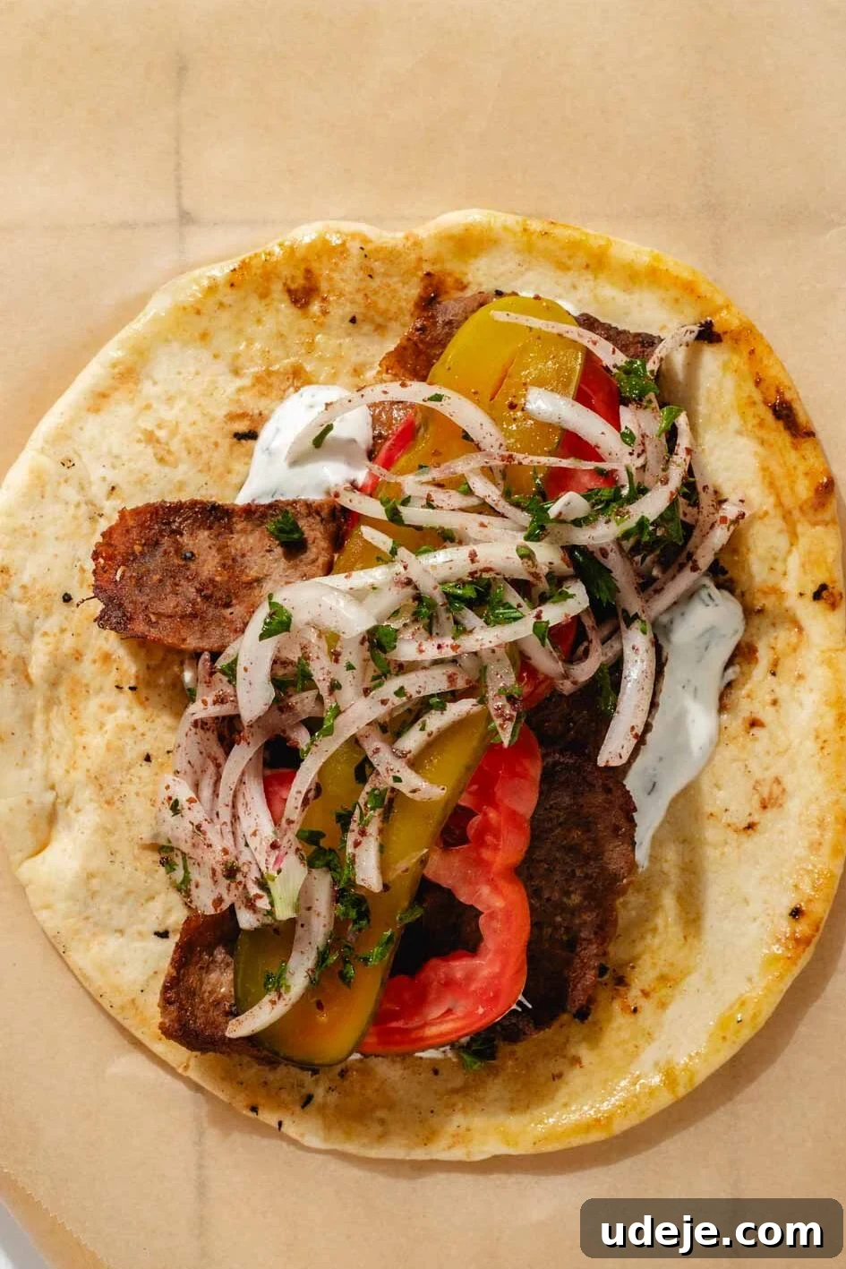 One-Pan Lamb and Beef Doner Kebab 19 A beautifully assembled pita bread, generously filled with succulent doner meat and fresh toppings including crisp pickles, vibrant sliced onions, and juicy tomato slices, rests on a sheet of brown parchment paper on a clean white tiled background, ready to be devoured.