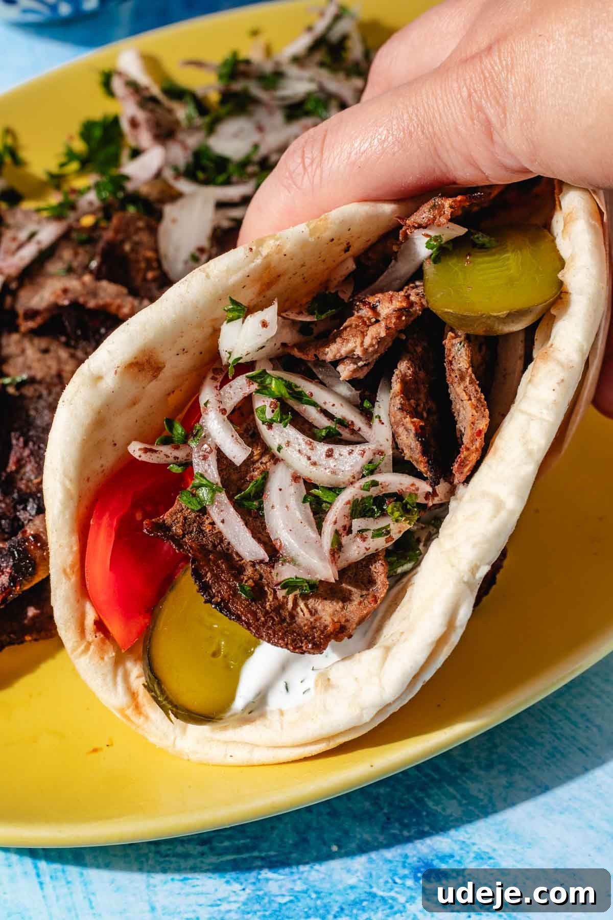 One-Pan Lamb and Beef Doner Kebab 20 A delectable doner sandwich is held delicately on a bright yellow platter, with more tantalizing doner slices and fresh onion slices artfully arranged on a striking blue background, inviting diners to indulge in this classic wrap.