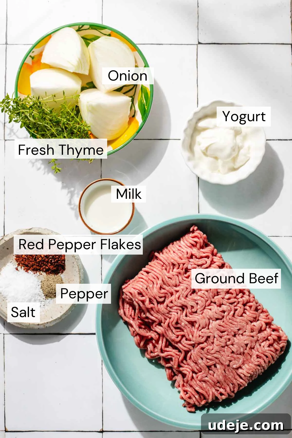 One-Pan Lamb and Beef Doner Kebab 3 A beautifully arranged ingredient shot for doner kebab, featuring quartered onion and fresh thyme in a yellow and green bowl. Beside it, small bowls hold creamy milk and rich Greek yogurt. A small plate displays a trio of vibrant red pepper flakes, freshly ground black pepper, and coarse salt. All ingredients are artfully placed around a blue bowl brimming with fresh ground beef on a clean white tiled background, highlighting the fresh components for this delicious dish.