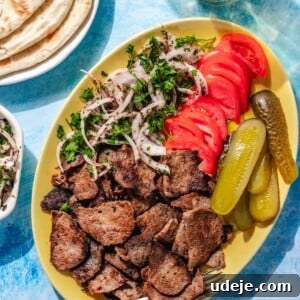 One-Pan Lamb and Beef Doner Kebab 21 Yellow platter with doner meat slices, sliced onions, sliced tomatoes, slice pickles, a fork on a blue background with pita on a plate in a corner and white doner sauce in another, and a glass of water