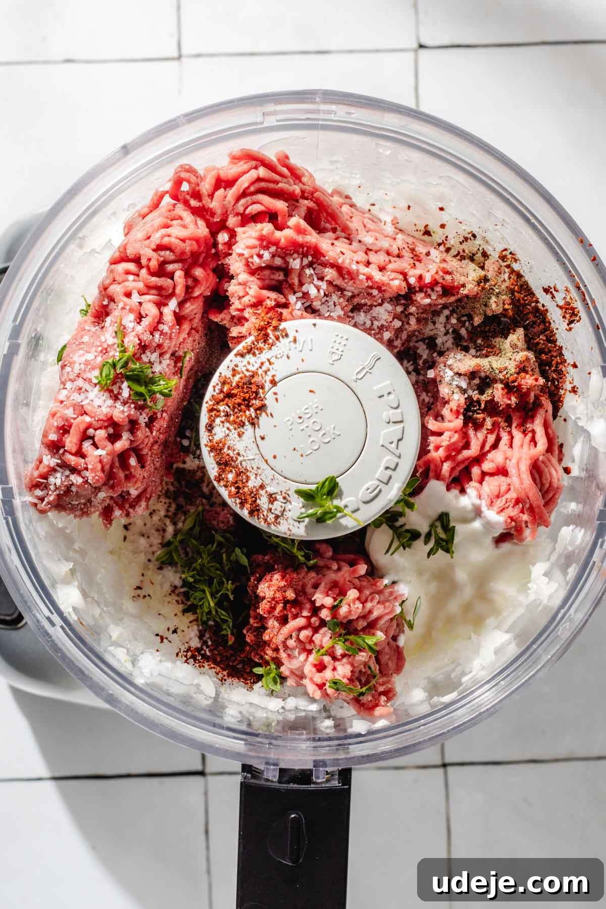 One-Pan Lamb and Beef Doner Kebab 7 Finely diced onions in a food processor, alongside generous portions of ground beef, fresh thyme sprigs, and a sprinkle of various seasonings, all resting on a clean white tiled background, preparing for the next step of blending.