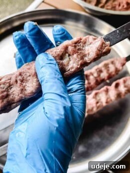 A hand wearing a blue glove shapes raw ground meat onto a skewer over a metal tray, preparing authentic Iraqi Kabab for grilling.