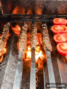 Four skewers of Iraqi Kabab grill over open flames next to sliced tomatoes on a barbecue grill.