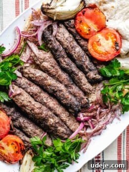 Iraqi Kabab grilled kebabs served on a white platter with roasted tomatoes, onions, fresh parsley, sliced red onions, and pita bread on the side. The plate sits on a striped tablecloth.