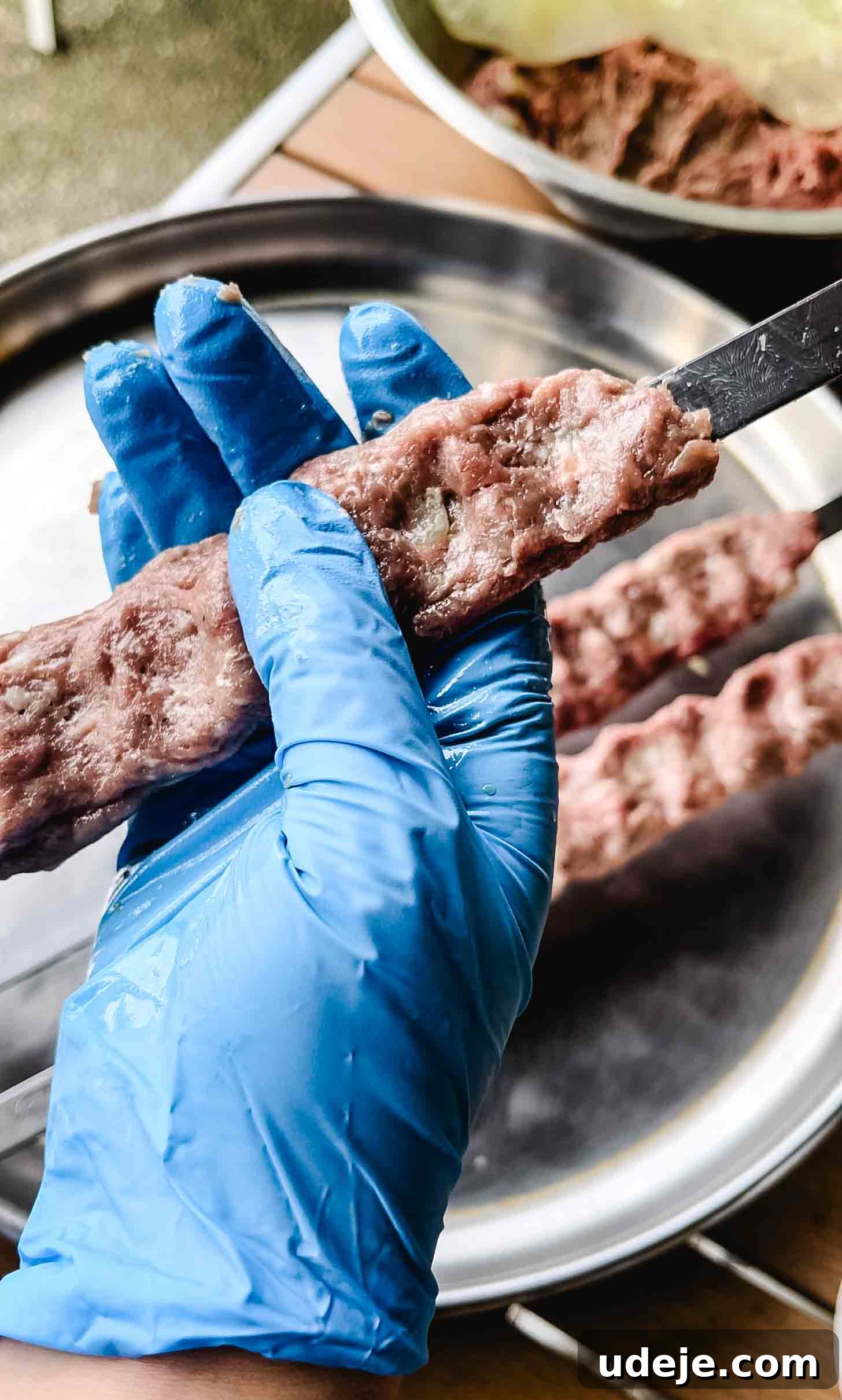 Sizzling Iraqi Kabab A Taste of Tradition 6 A hand wearing a blue disposable glove shapes raw ground meat onto a flat metal skewer, preparing authentic Iraqi Kabab for grilling. The meat is pressed and elongated along the skewer.