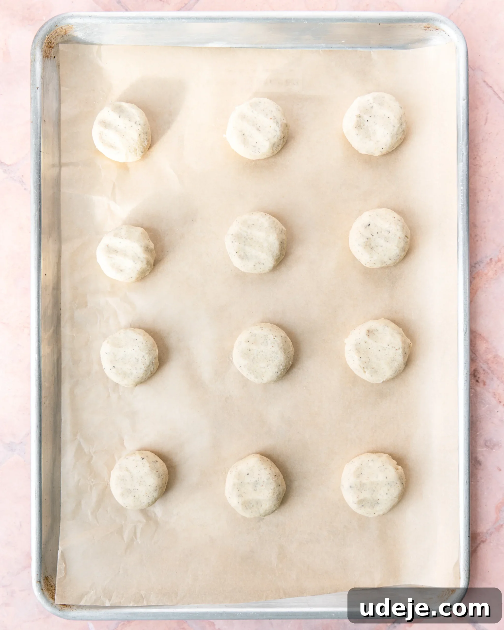 Fragrant Cardamom Shortbread Cookies 12 Baking sheet lined with parchment with 12 evenly spaced dough balls that have been lightly flattened on a light red tiled background.