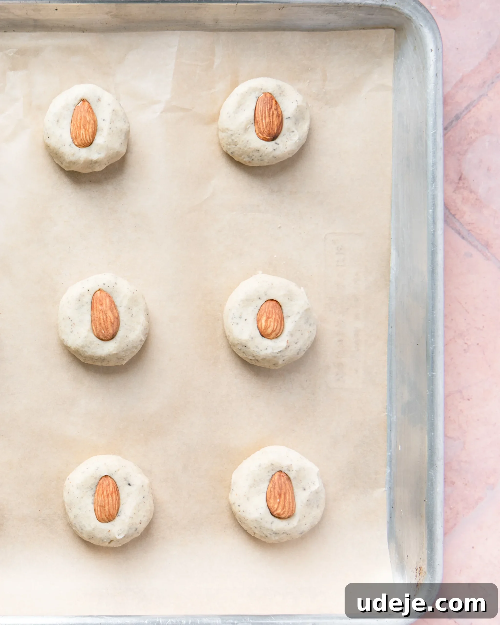 Fragrant Cardamom Shortbread Cookies 13 Baking sheet lined with parchment with 12 evenly spaced dough balls that have been lightly flattened with an almond in the center on a light red tiled background.