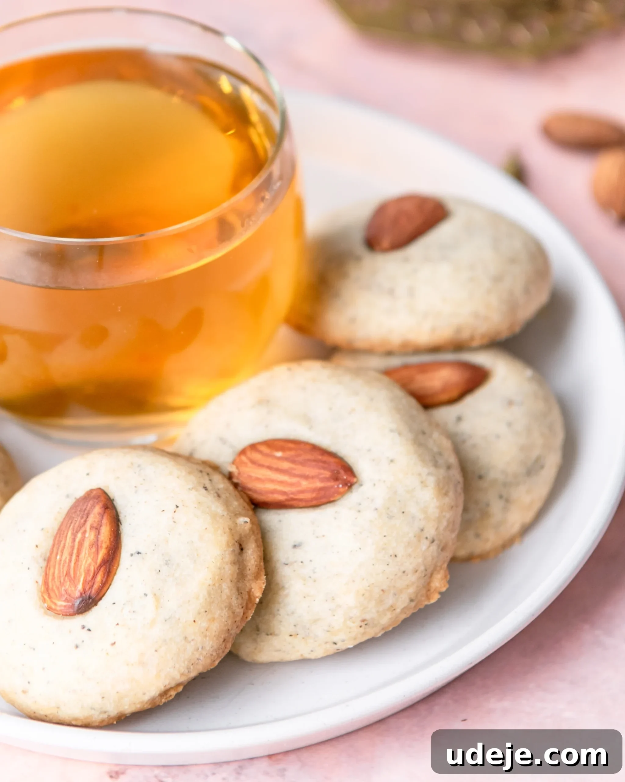 Fragrant Cardamom Shortbread Cookies 15 Round cookies with a whole almond in the centre on a white plate with a glass cup of herbal tea on a light red tiled background.