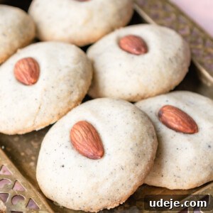 Round lightly coloured cookies with a whole almond in the centre on a gold coloured tray on a light red tiled background.