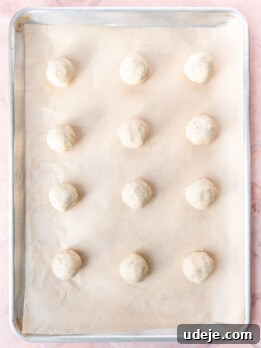 Baking sheet lined with parchment with 12 evenly spaced dough balls on a light red tiled background.