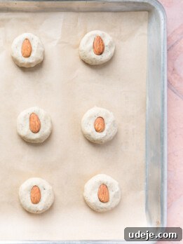 Baking sheet lined with parchment with 12 evenly spaced dough balls that have been lightly flattened with an almond in the center on a light red tiled background.