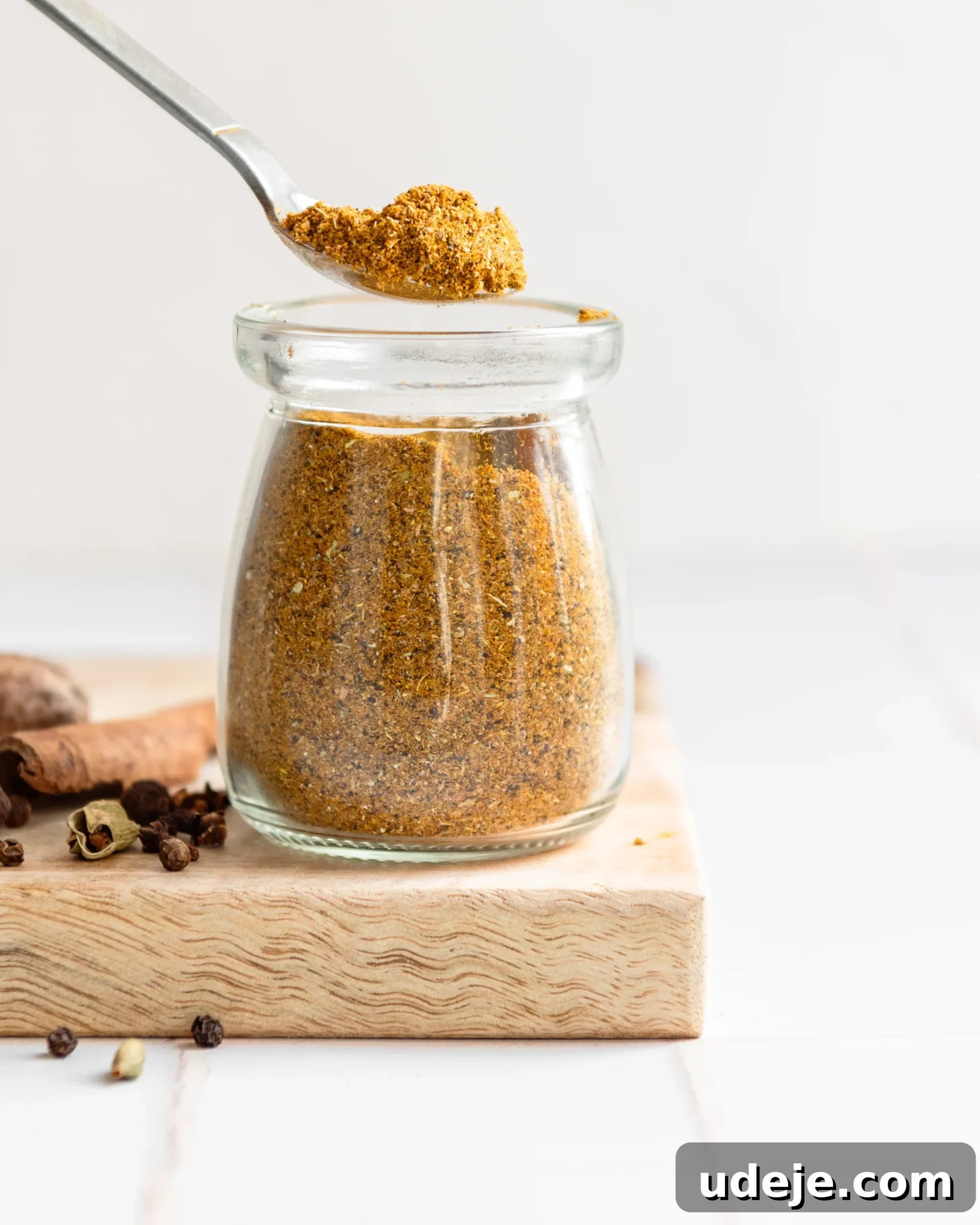 Handcrafted Baharat Mix 2 A small glass jar filled with homemade Baharat spice blend, with a metal spoon resting inside. Surrounding the jar are various whole spices like peppercorns, cinnamon sticks, and star anise, all arranged on a rustic wooden cutting board against a white tiled background.