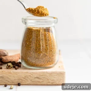 A small glass jar filled with homemade Baharat spice blend, with a metal spoon resting inside. Surrounding the jar are various whole spices like peppercorns, cinnamon sticks, and star anise, all arranged on a rustic wooden cutting board against a white tiled background.