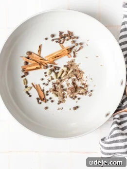whole cinnamon sticks, cardamom, allspice, nutmeg, cumin seeds, cloves in a light grey pan with a black striped tea towel on a white tiled background