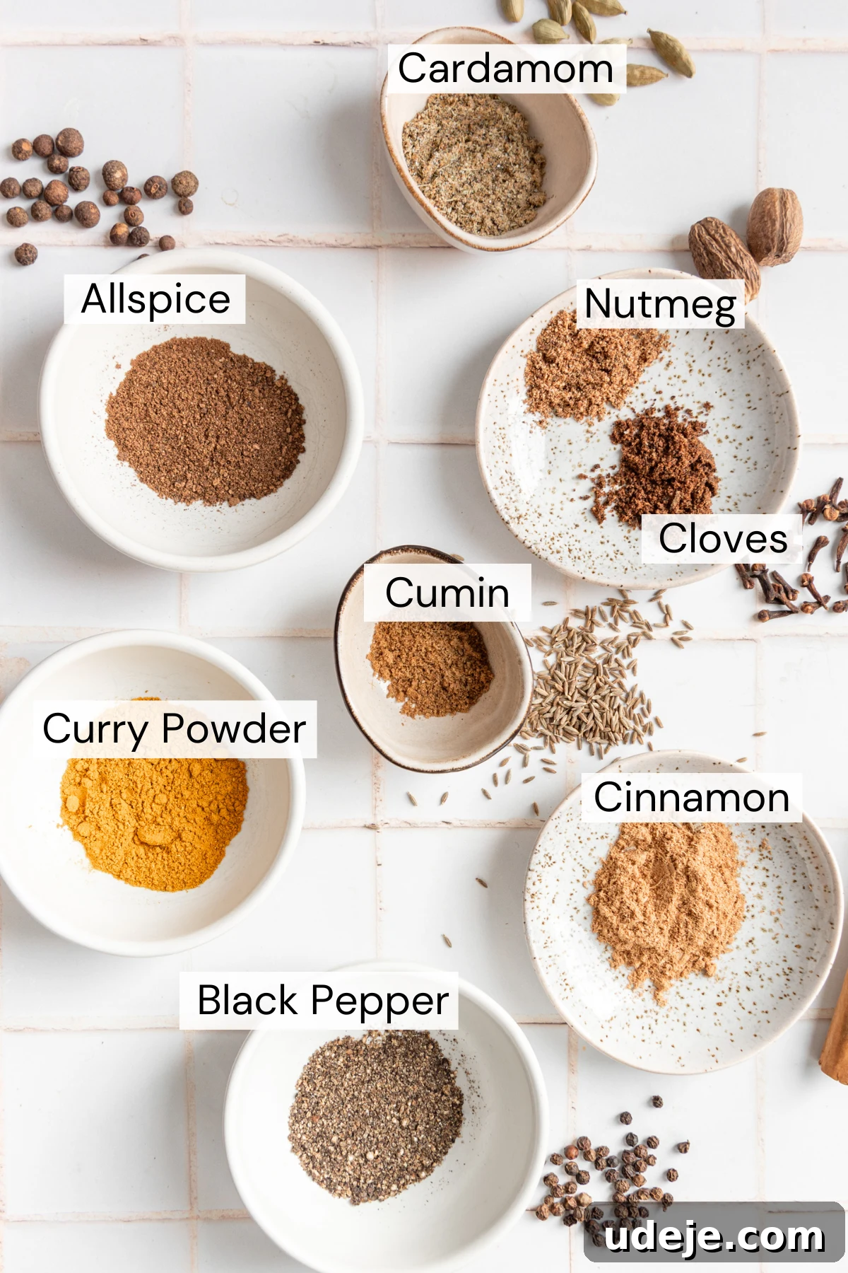 Handcrafted Baharat Mix 3 A vibrant collection of Baharat ingredients, showcasing small bowls of ground cardamom, allspice, nutmeg, cloves, cumin, curry powder, cinnamon, and black pepper, each accompanied by its whole spice counterpart, arranged artfully on a white tiled background.