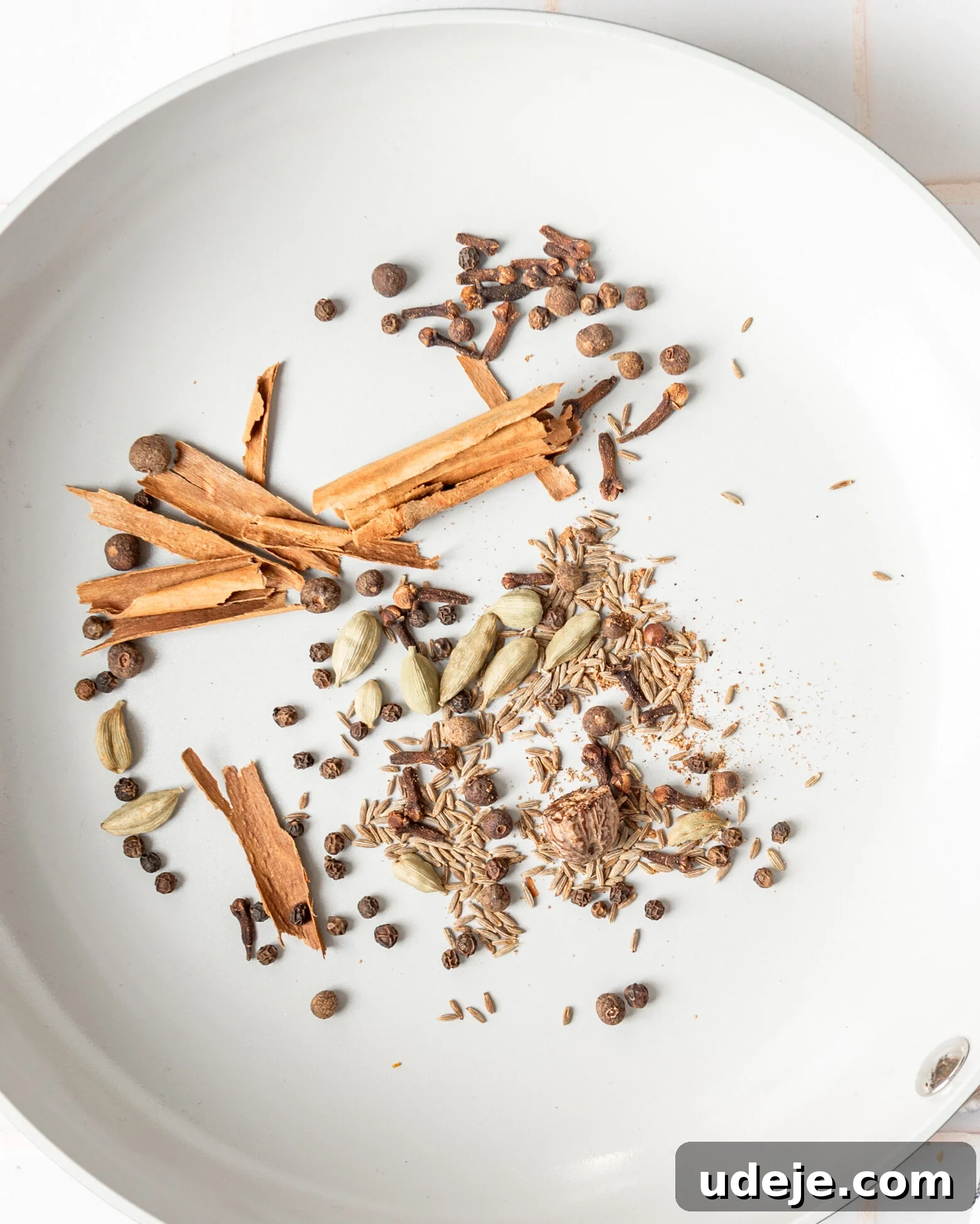 Handcrafted Baharat Mix 4 A selection of whole spices including cinnamon sticks, cardamom pods, allspice berries, nutmeg, cumin seeds, and cloves gently toasting in a light grey pan, with a black striped tea towel beside it on a white tiled background.