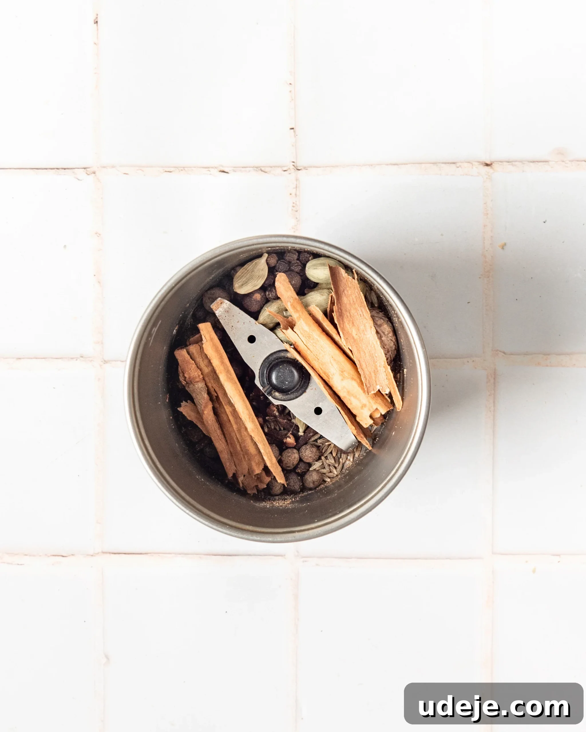 Handcrafted Baharat Mix 5 Whole spices, including cinnamon sticks, cardamom, allspice, nutmeg, cumin seeds, and cloves, being loaded into a spice grinder on a white tiled background, ready for grinding.