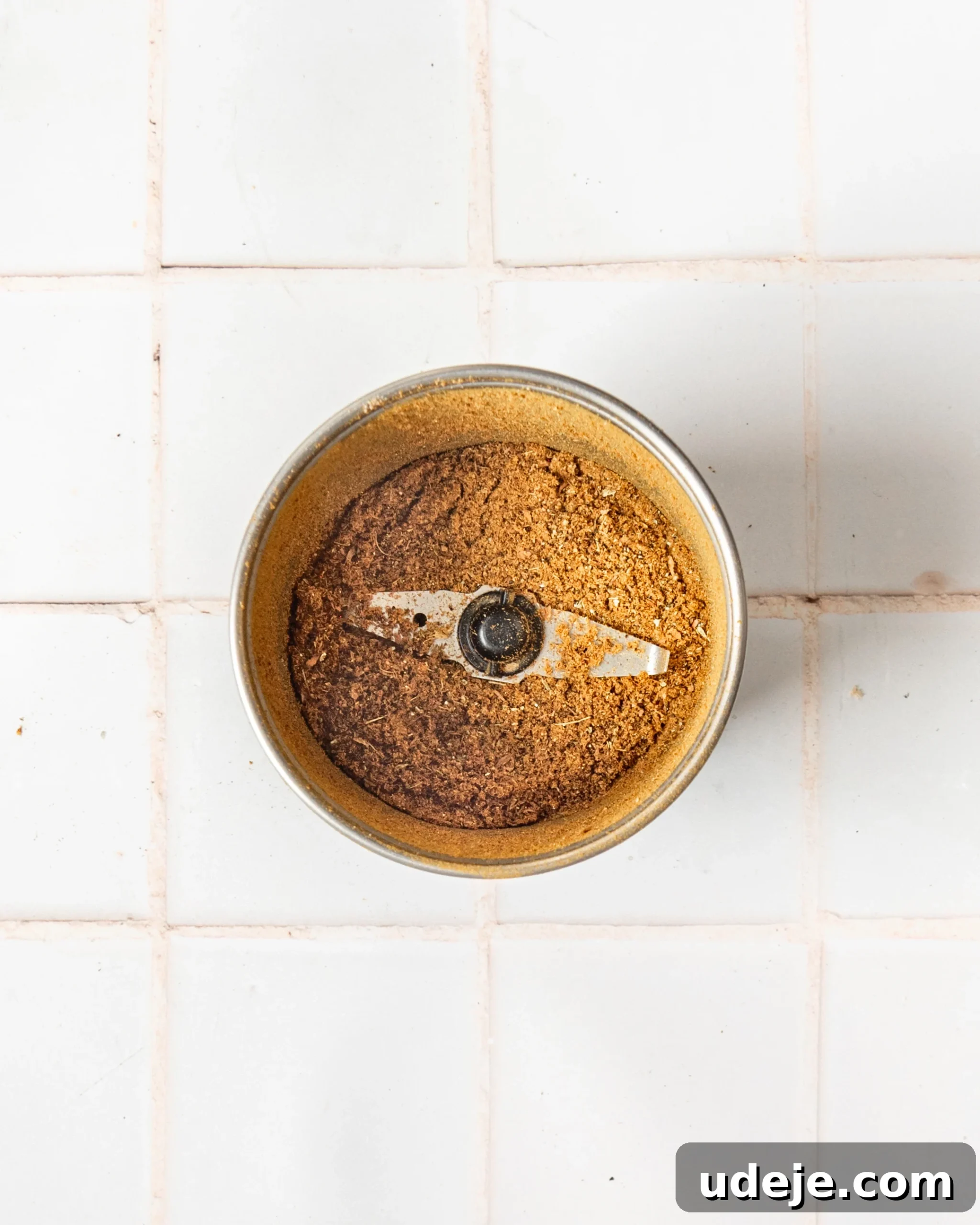 Handcrafted Baharat Mix 6 Freshly ground Baharat spice blend filling the basin of a spice grinder, showcasing its rich color and fine texture, set against a clean white tiled background.