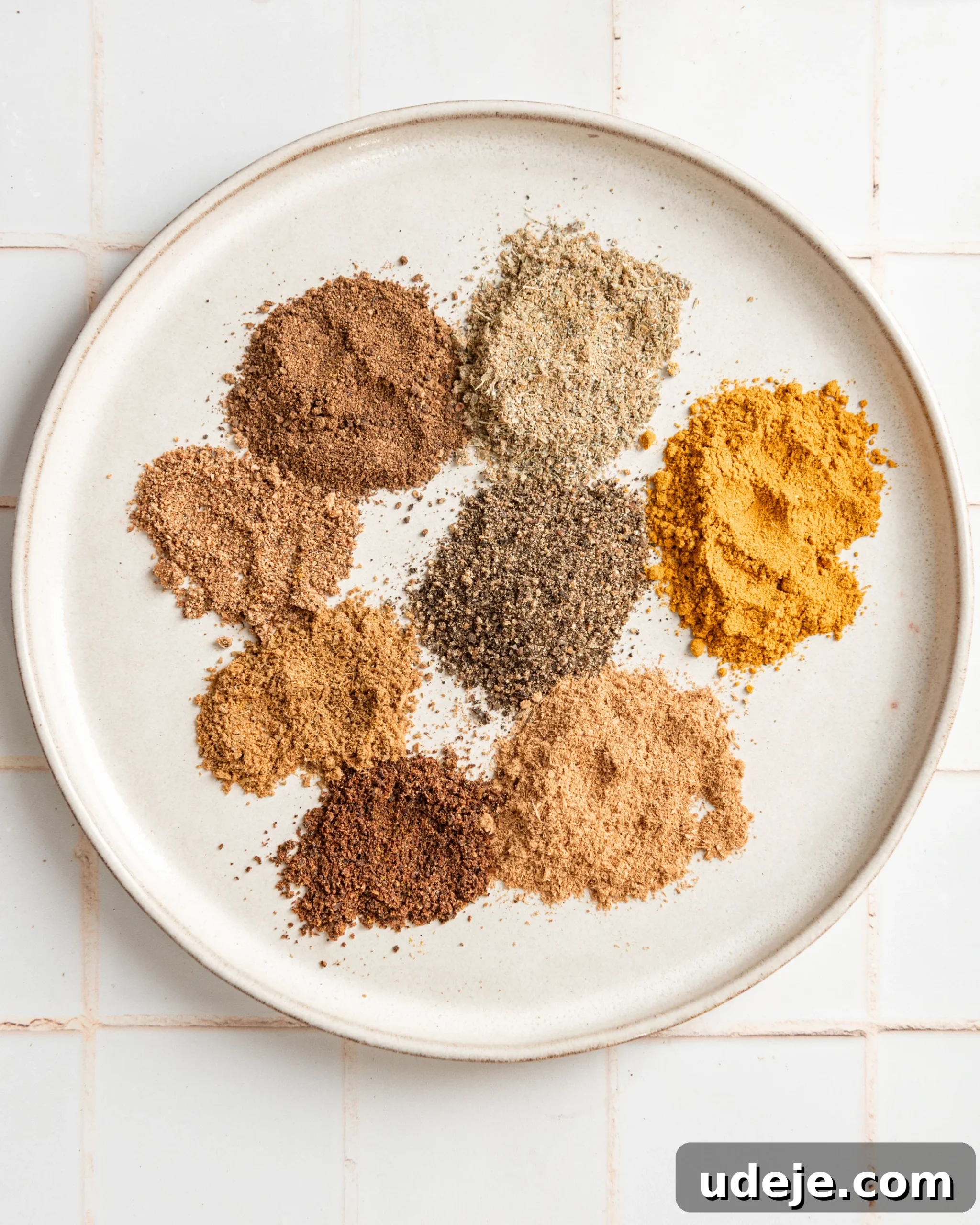 Handcrafted Baharat Mix 7 An array of distinct ground spices for Baharat carefully arranged on a white plate, ready for mixing, against a white tiled background.