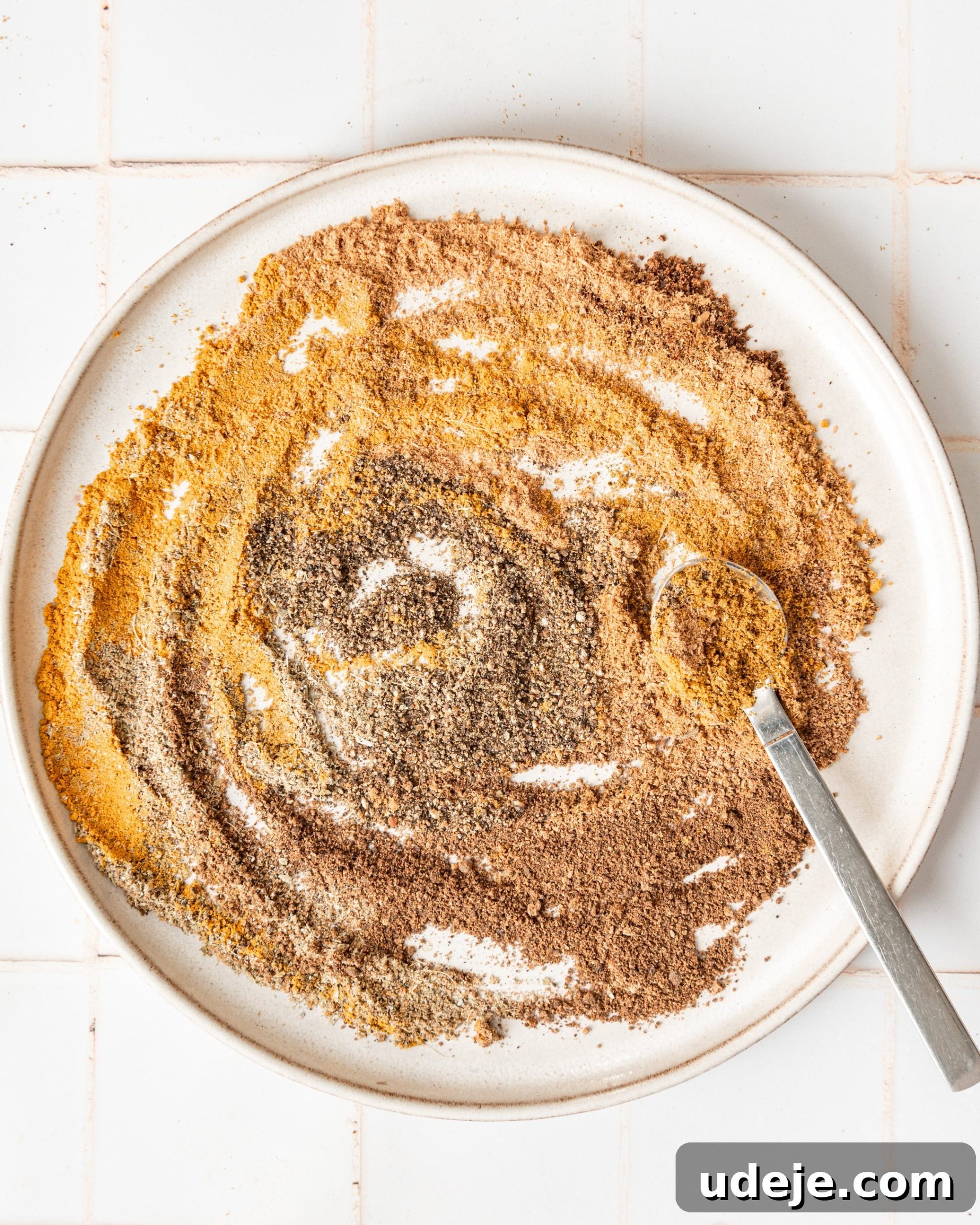 Handcrafted Baharat Mix 8 A metal spoon gently mixing the assorted ground Baharat spices on a white plate, ensuring a uniform blend, all on a white tiled background.