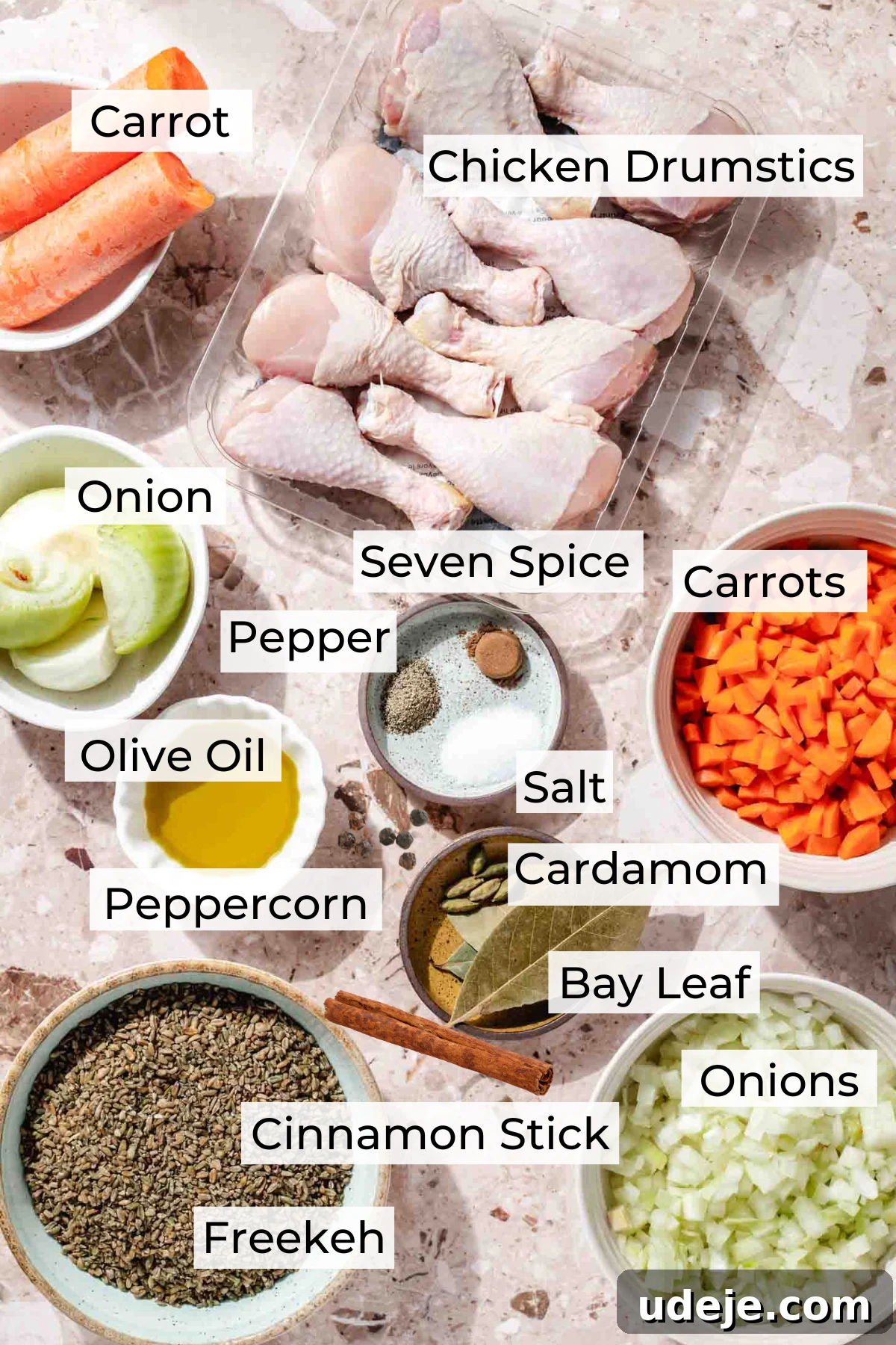 Slow-Simmered Chicken Freekeh Soup 3 A vibrant array of fresh ingredients laid out on a terrazzo background. Chicken drumsticks are in a clear container, alongside whole carrots, halved onions, and chopped carrots in white bowls. Olive oil, a small plate of seven spice, pepper, and salt, and another bowl with cardamom pods, bay leaves, a cinnamon stick, and peppercorns complete the spice selection. A bowl of freekeh and chopped onions are also visible, showcasing the fresh components ready for soup preparation.
