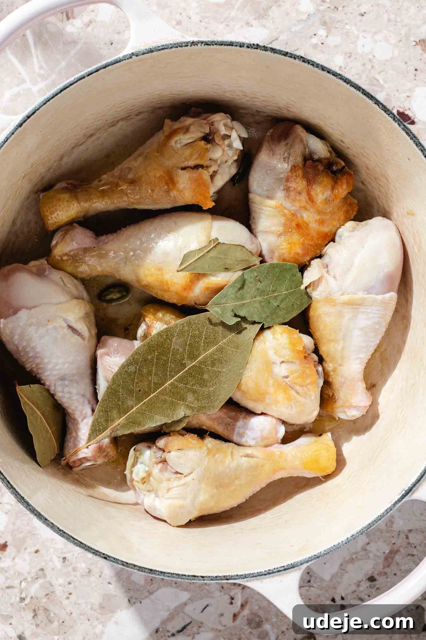 Slow-Simmered Chicken Freekeh Soup 4 Browned chicken legs nestled with bay leaves in a white soup pot, resting on a terrazzo background, ready for the next step in broth preparation.