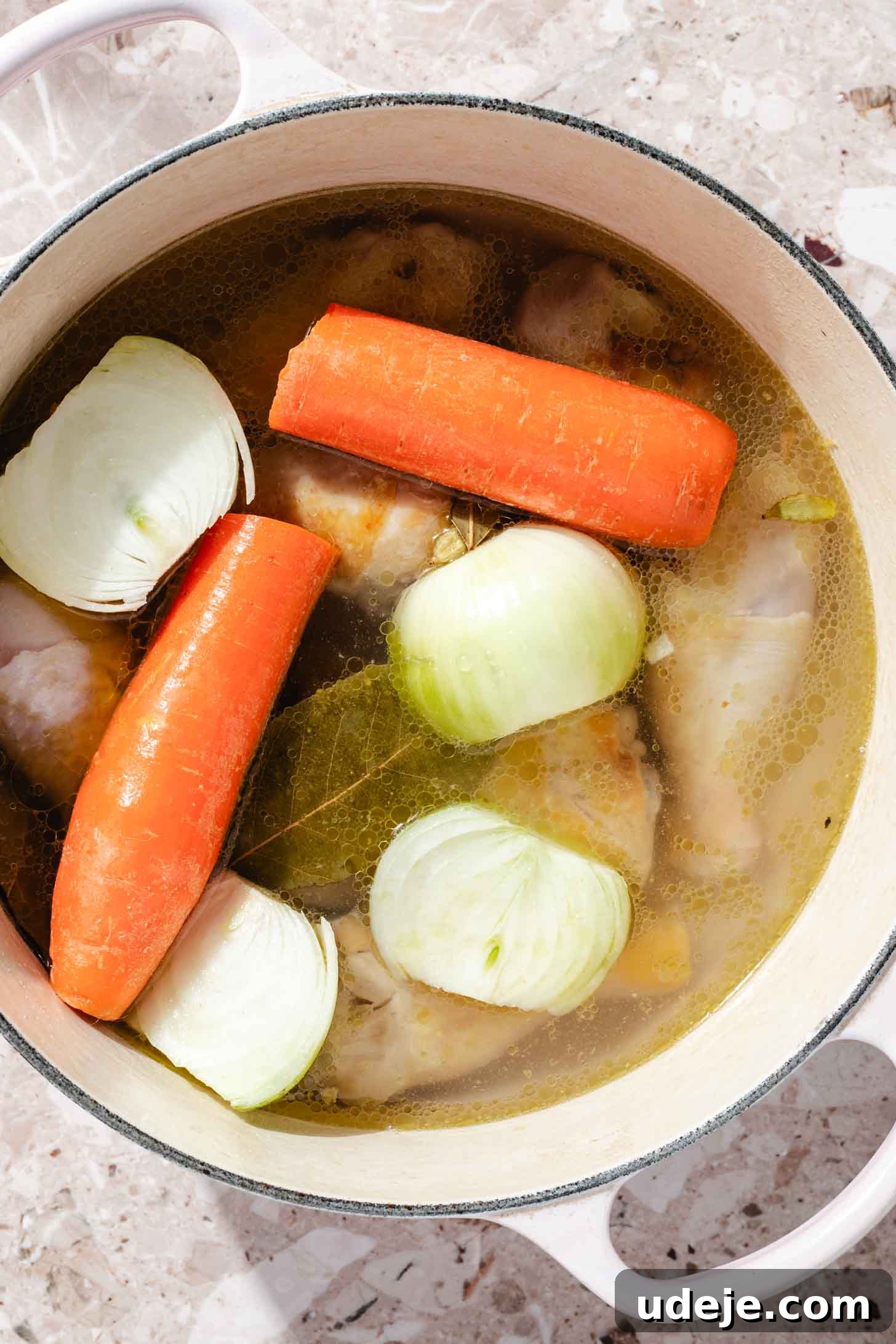 Slow-Simmered Chicken Freekeh Soup 5 Chicken legs, whole carrots, halved onions, and bay leaves are gently simmering in a flavorful broth within a white pot, placed on a terrazzo background, showcasing the ongoing broth preparation.