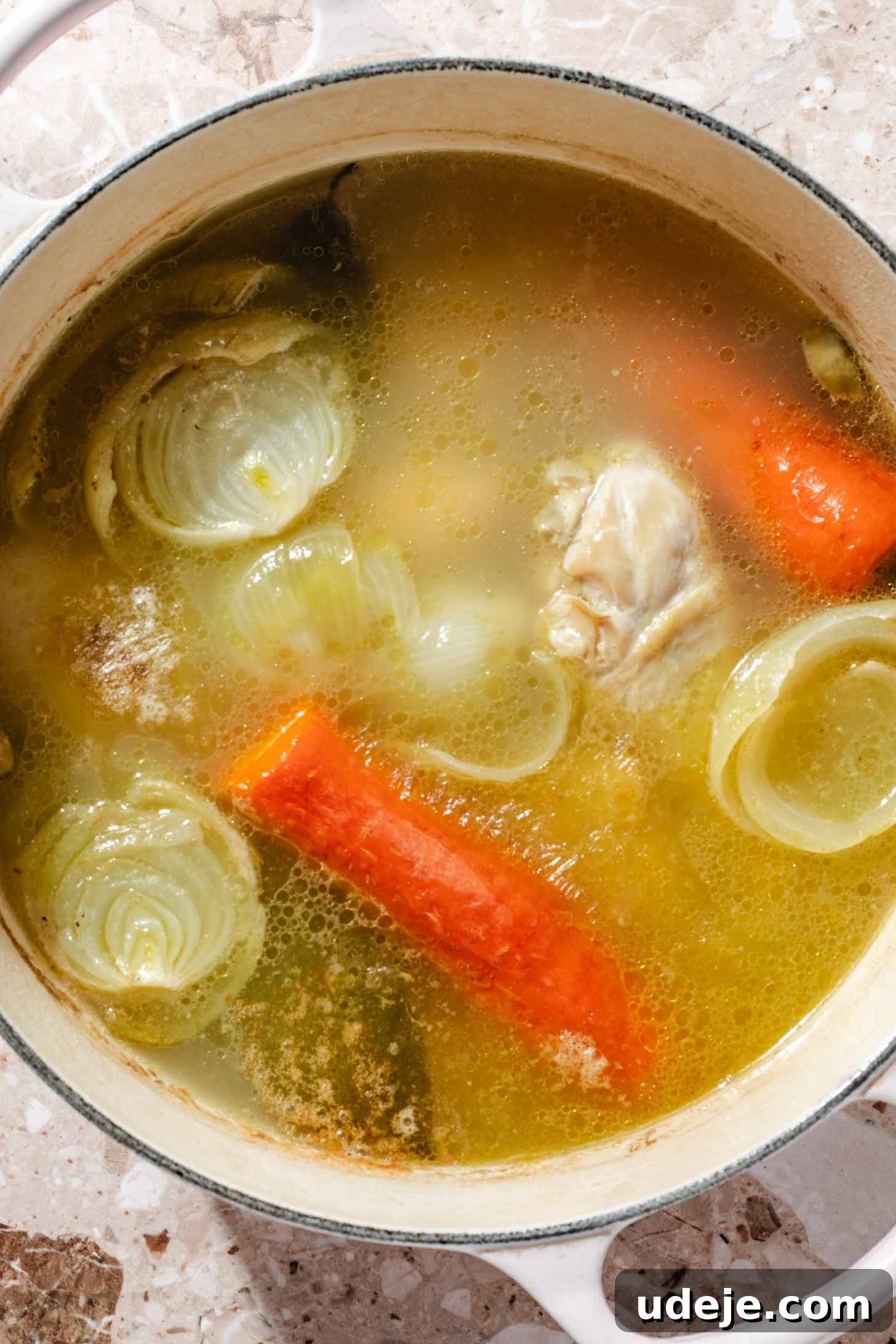 Slow-Simmered Chicken Freekeh Soup 6 Chicken legs, whole carrots, halved onions, and bay leaves are gently simmering in a flavorful broth within a white pot, placed on a terrazzo background, showcasing the ongoing broth preparation.