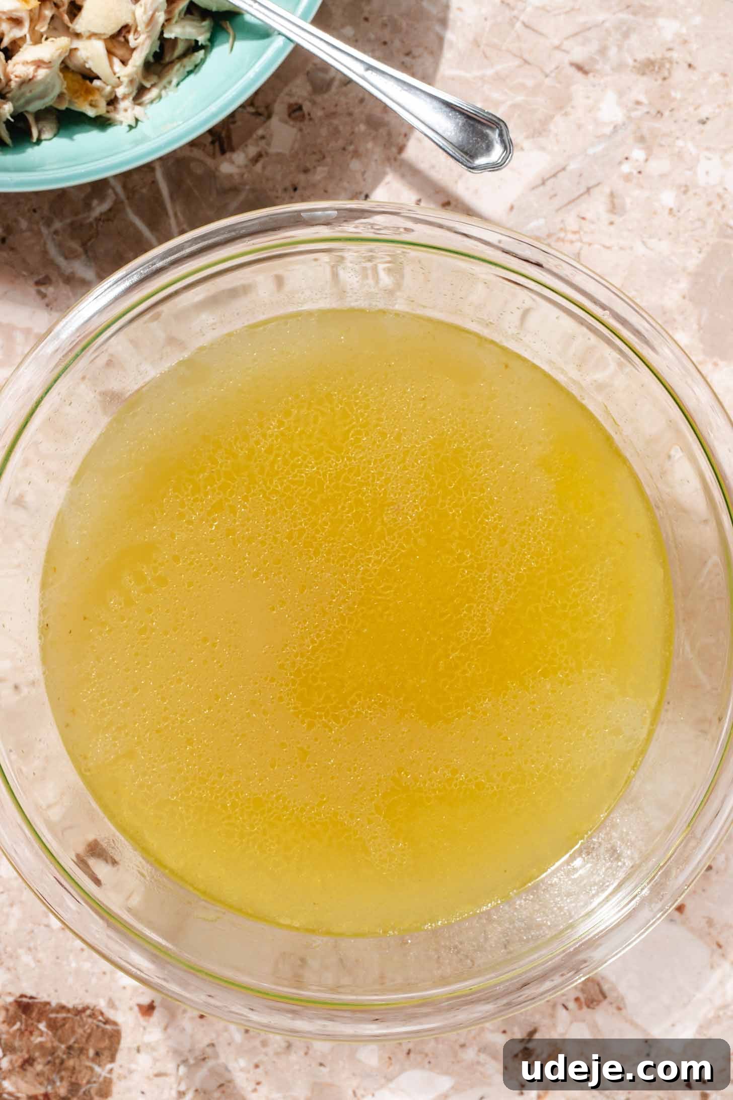 Slow-Simmered Chicken Freekeh Soup 7 Clear chicken broth is presented in a glass bowl, with a blue bowl of freshly shredded chicken positioned above it, accompanied by a spoon, all set on a terrazzo background, ready for soup assembly.