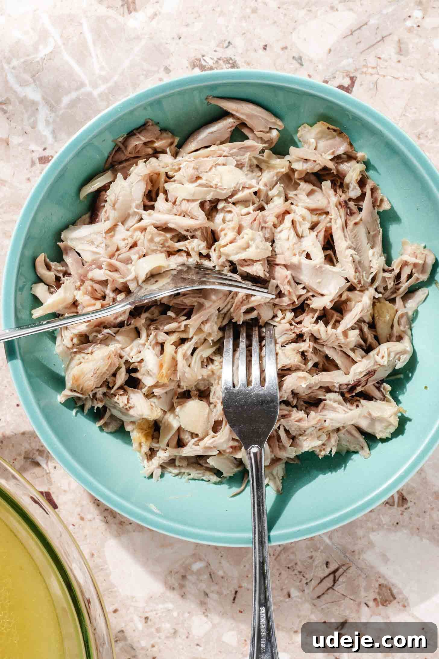 Slow-Simmered Chicken Freekeh Soup 8 A blue bowl holds tender shredded chicken with two metal forks, alongside a glass bowl of golden chicken broth, all arranged on a terrazzo background, ready to be combined into a delicious soup.