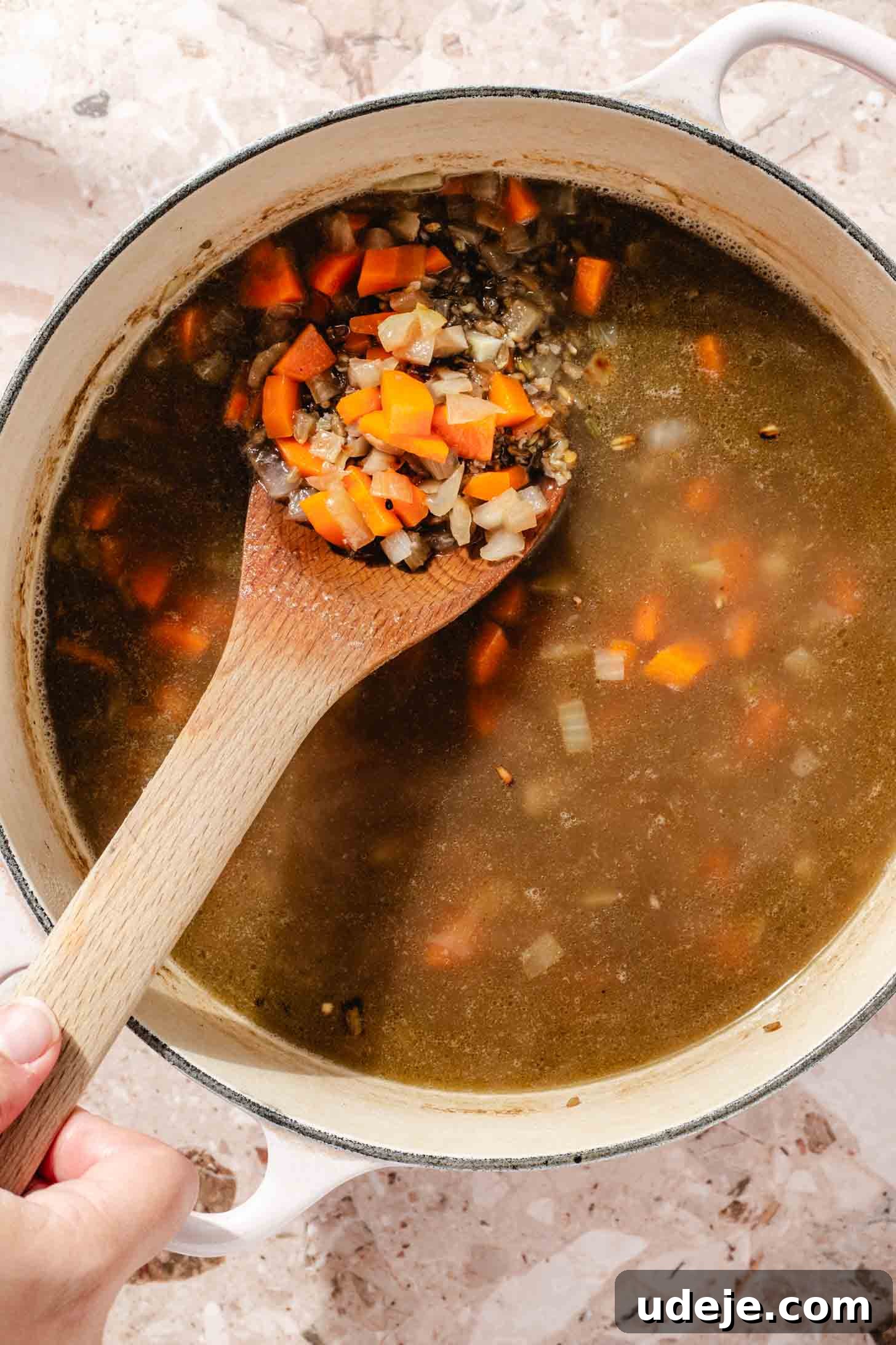 Slow-Simmered Chicken Freekeh Soup 9 A large white pot holds a comforting soup, rich with freekeh, chopped carrots, and onions in a savory broth, with a wooden spoon resting inside, all on a terrazzo background.