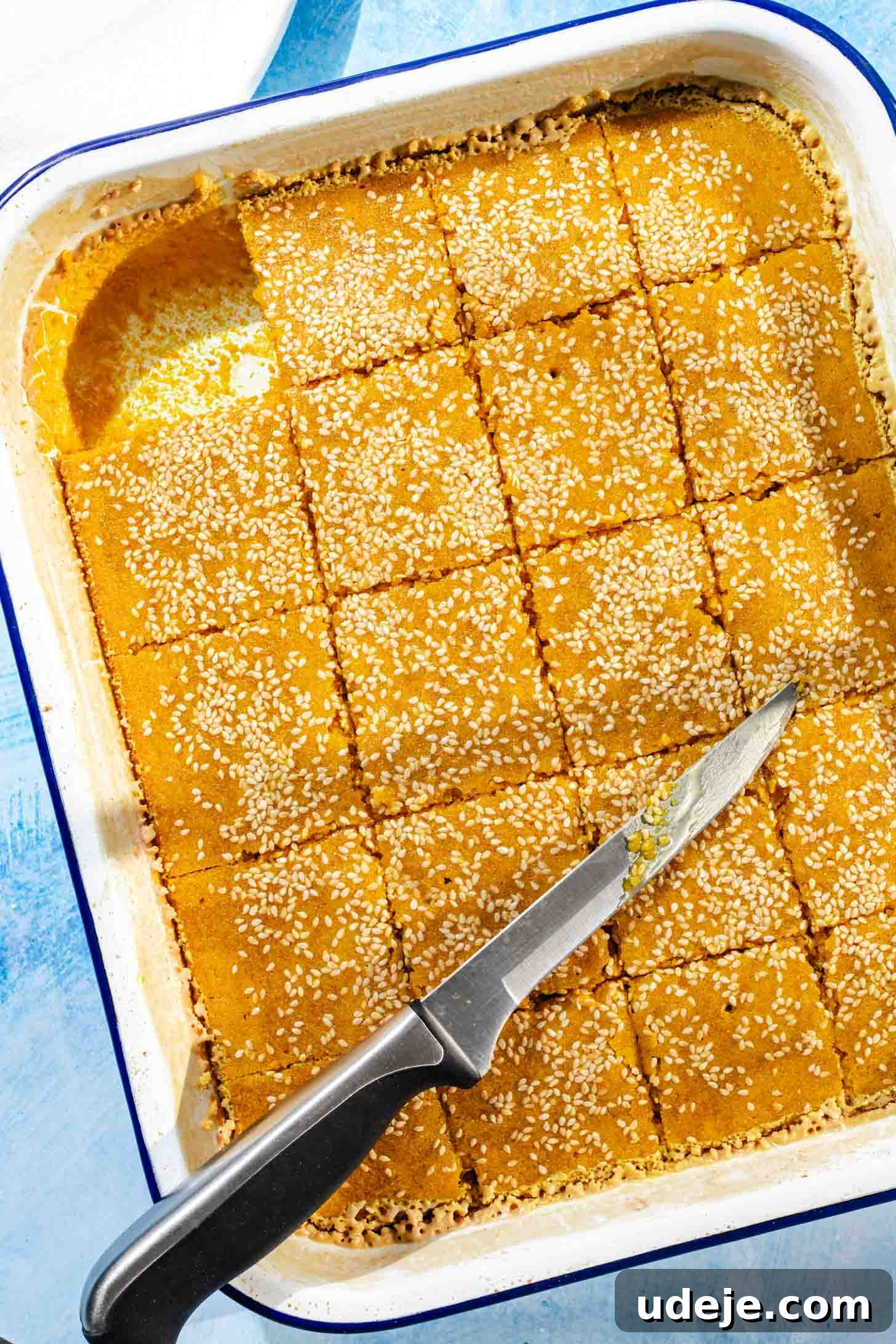 Aromatic Golden Semolina Cake 11 Baked Sfouf yellow cake in a white rectangular baking pan with a blue rim, neatly cut into squares with one piece removed, a knife resting on the side, all on a blue background.