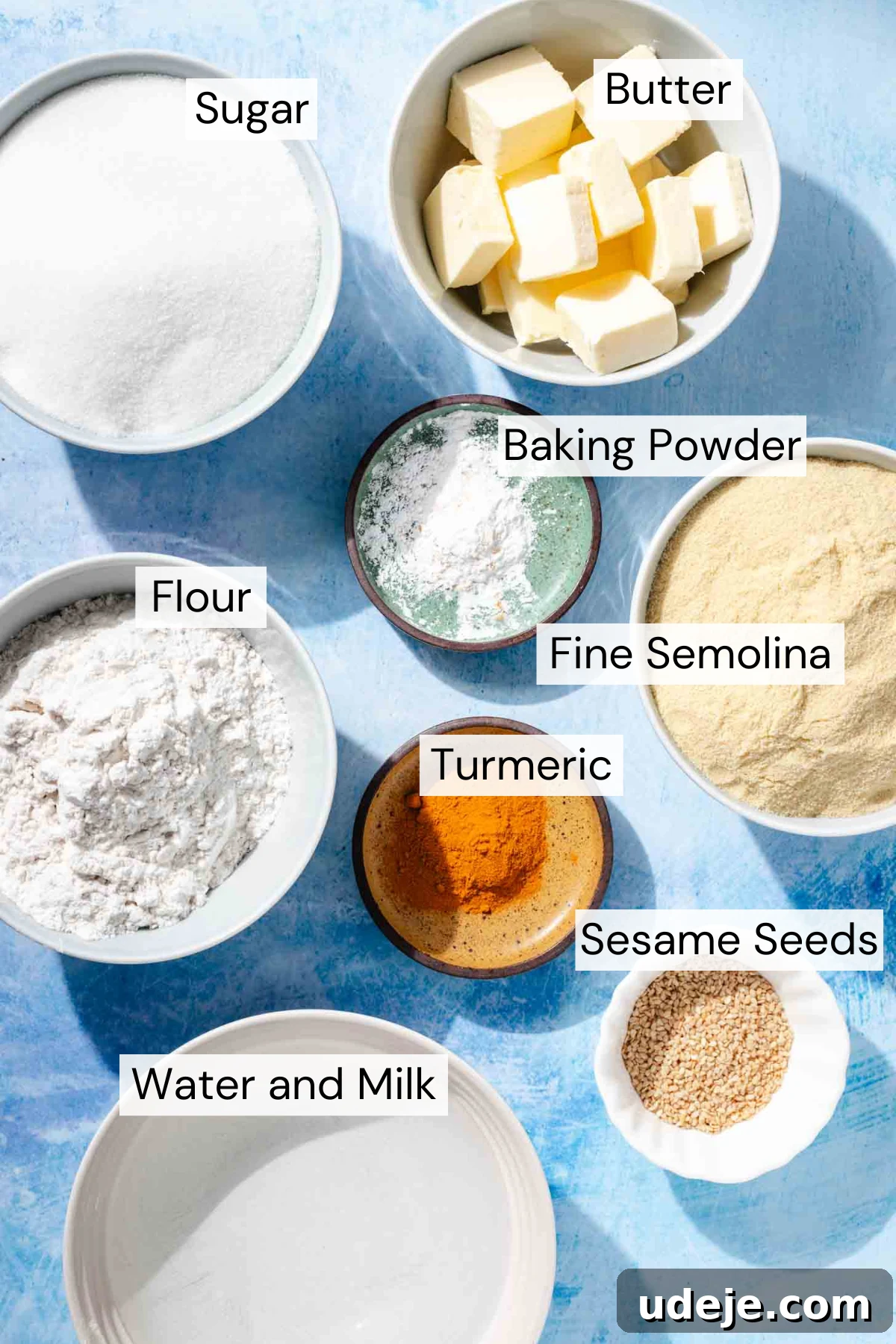 Aromatic Golden Semolina Cake 3 A flat lay of all Sfouf ingredients: cubed butter, sugar, baking powder, fine semolina, all-purpose flour, turmeric, sesame seeds, water, and milk, all presented in white bowls and small plates on a vibrant blue background.