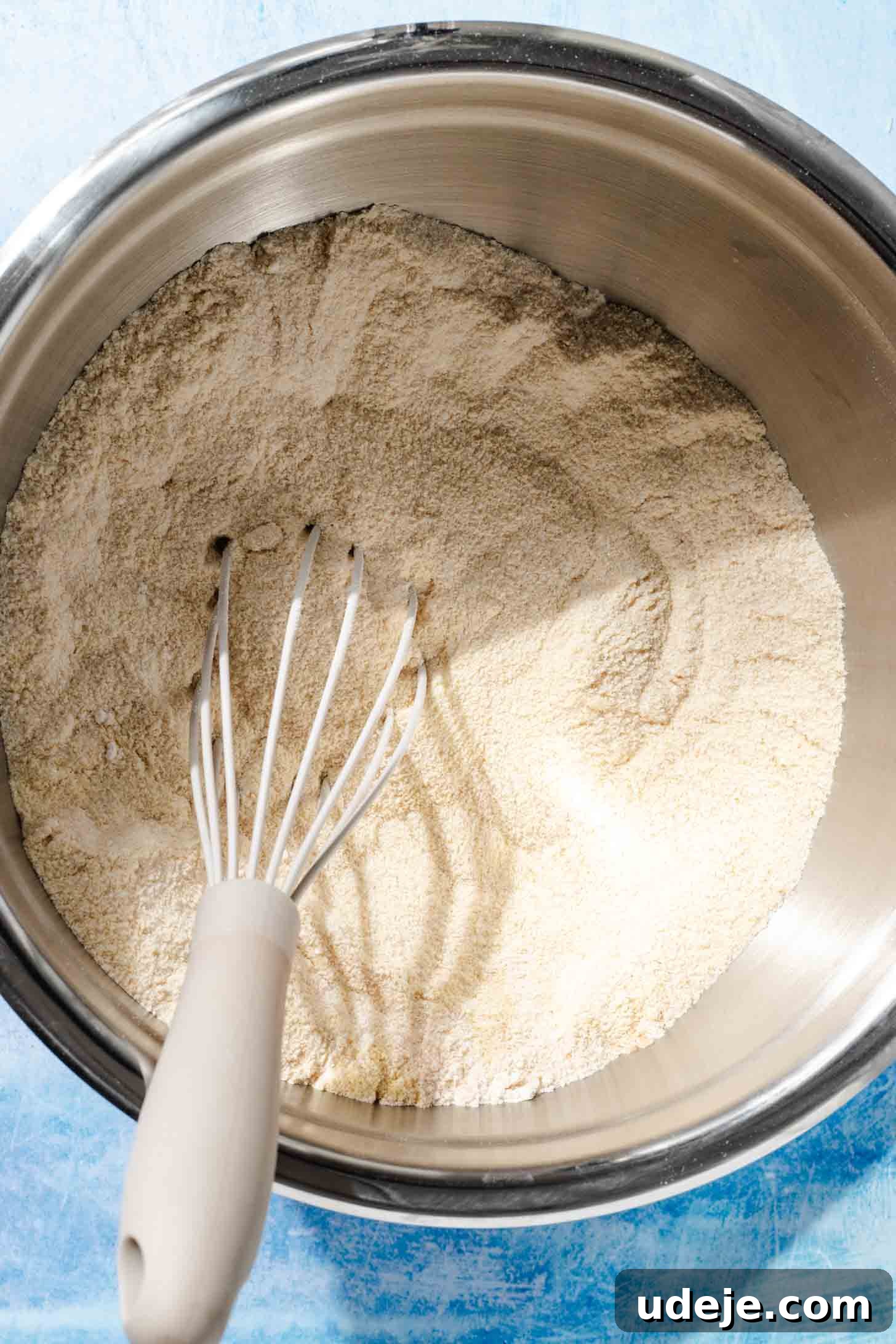 Aromatic Golden Semolina Cake 4 A metal mixing bowl containing white flour and fine semolina, with a whisk resting inside, all set on a blue background.