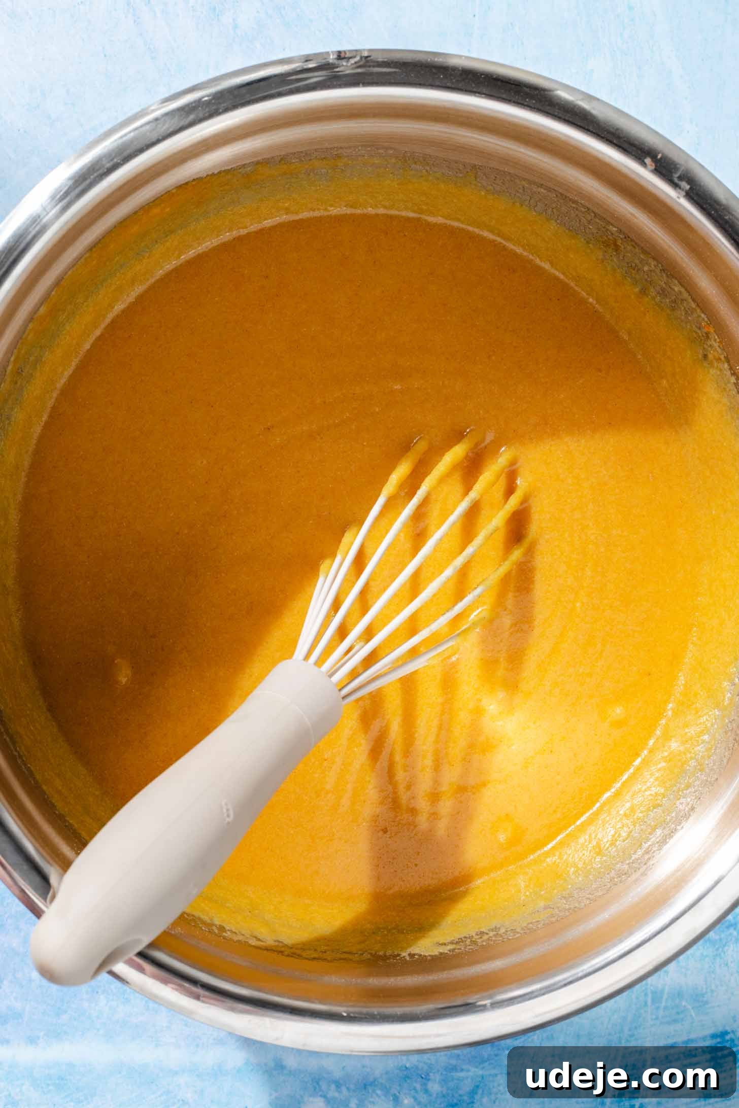 Aromatic Golden Semolina Cake 5 Vibrant yellow-colored Sfouf batter in a metal mixing bowl with a whisk, resting on a blue background.